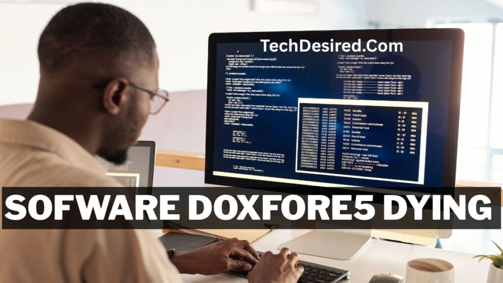 A person sitting at a desk and working on a computer displaying lines of code on a large monitor. The screen shows programming text and a data table, with the website name “TechDesired.com” at the top. A bold black banner across the bottom contains the text “sofware doxfore5 dying.” A keyboard, coffee cup, and other desk items are visible in the workspace.